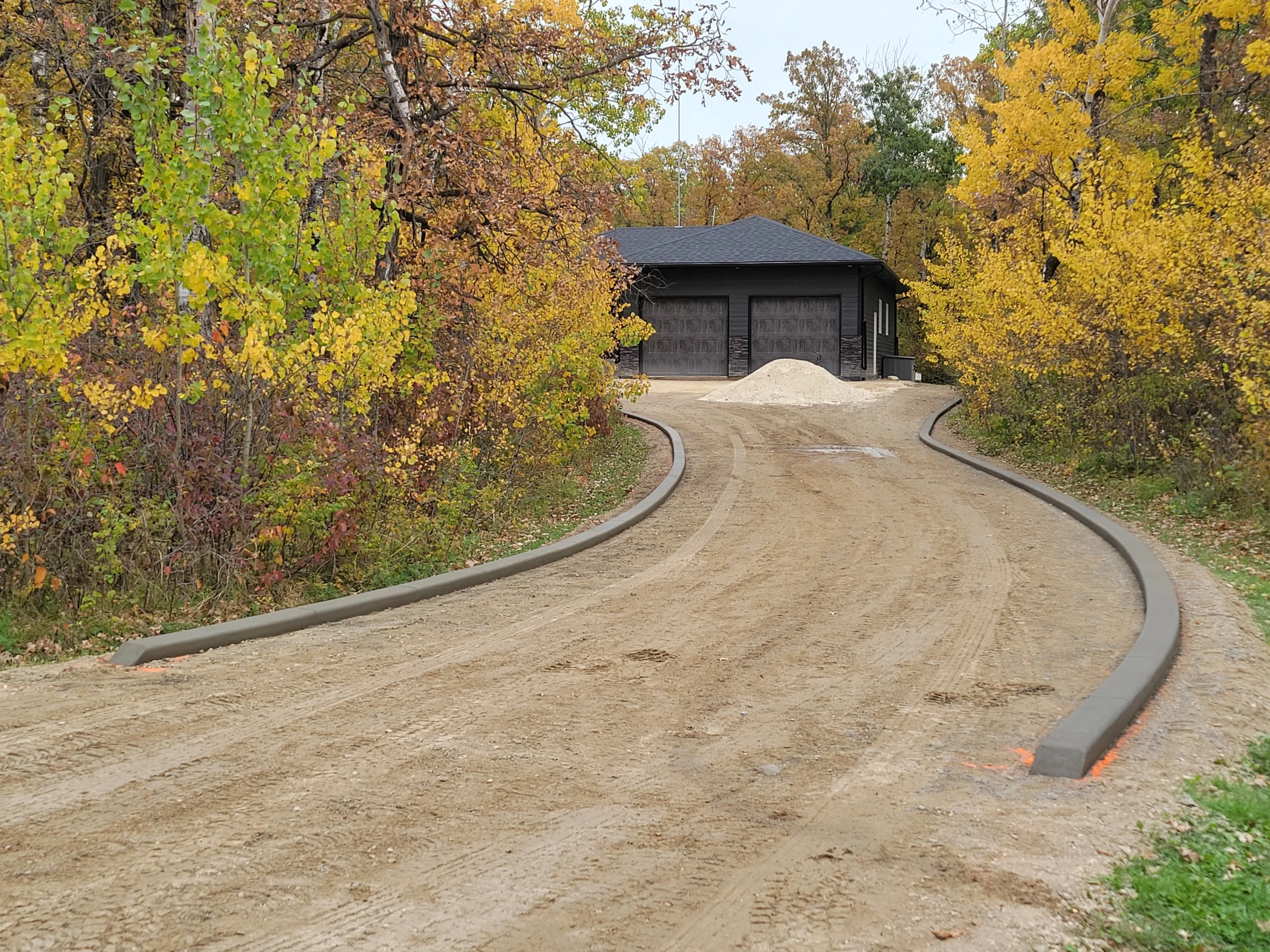 Long sweeping runs of concrete border edge curbing lining both sides of a rural driveway through autumn trees, installed by Maple Curbing.