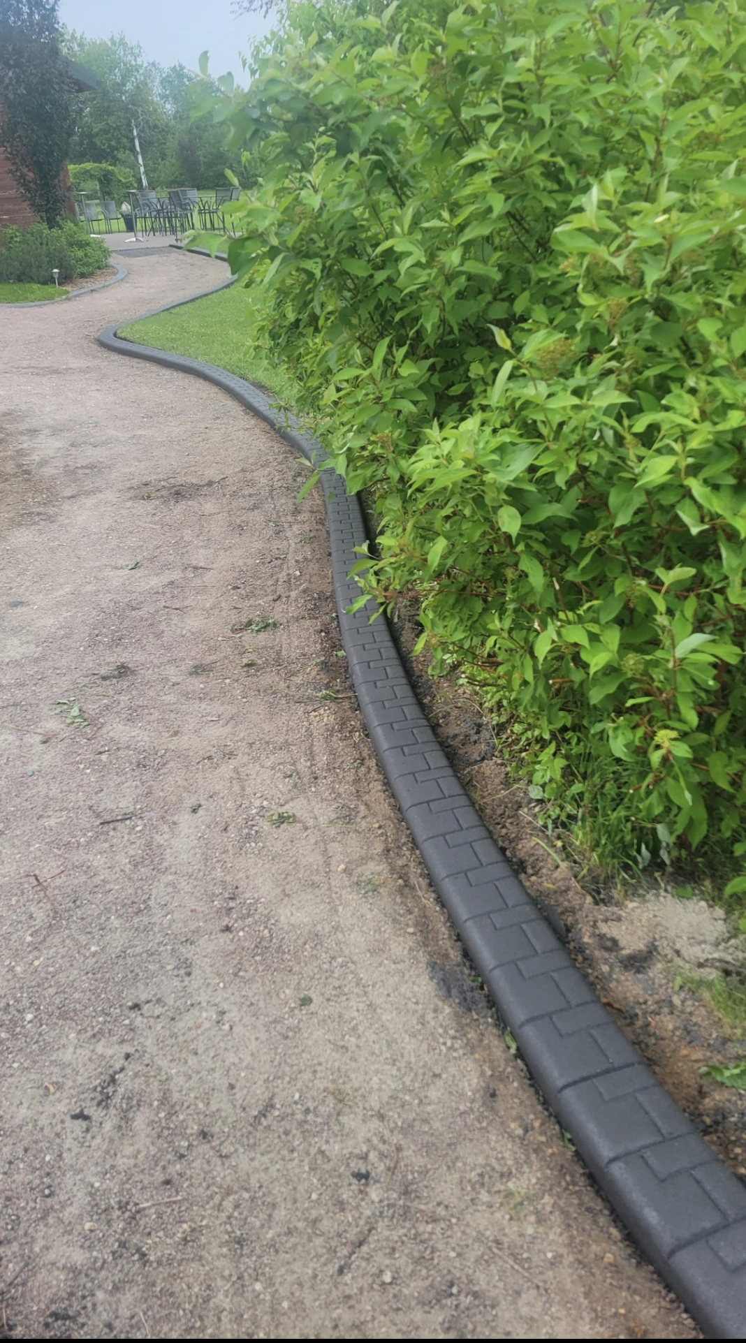 Smooth curved black flagstaff concrete curbing bordering a gravel driveway amid green shrubs and trees in a residential landscape, installed by Maple Curbing.