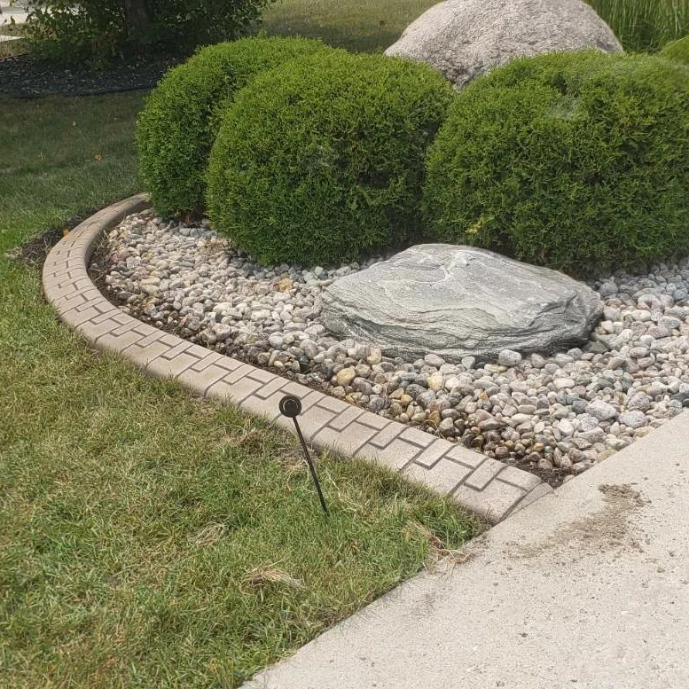 Brick-pattern flagstaff concrete curbing edging a landscaped rock garden with large boulders, rounded shrubs, and grass in a Manitoba yard, installed by Maple Curbing.