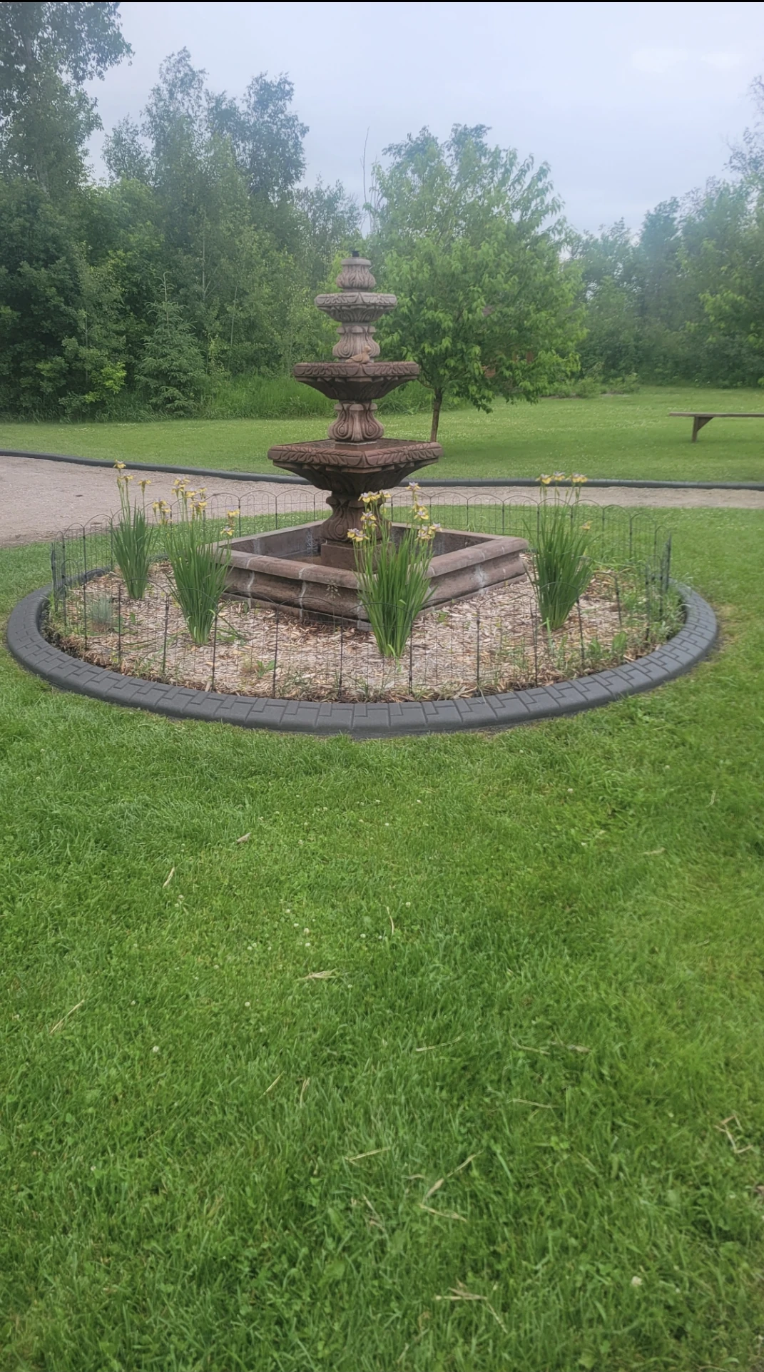 Circular flagstaff stamped concrete curbing surrounding a tiered stone fountain with ornamental grasses and mulch in a lush green lawn, installed by Maple Curbing.